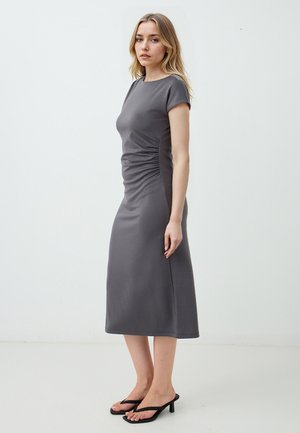 Graues Jerseykleid mit kurzen Ärmeln, figurbetonter Silhouette, seitlicher Raffung und knielangem Saum. Das Model trägt schwarze Sandalen.