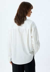 Hvid cotton skjorte med lange ærmer, rygsøm og buet kant. Har knapmanchetter og en afslappet pasform. Enkel tekstur, minimalistisk design.