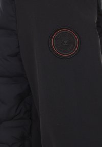 Giacca sportiva nera caratterizzata da una texture liscia e opaca, con una toppa circolare del logo in nero e arancione sulla manica.