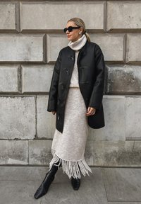 Schwarzer Ledermantel mit Riegelverschluss, crèmefarbener Strickpullover mit Stehkragen, fransiger beiger Rock und schwarze spitze Stiefel vor einer strukturierten Wand.