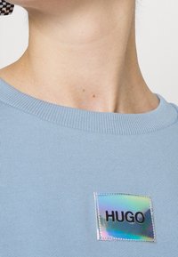 Collo e parte superiore del petto che indossano una camicia di tessuto a costine di un blu chiaro con un patch olografico etichettato "HUGO" sul lato sinistro.