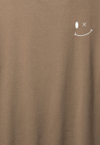 Tela marrón con un pequeño gráfico de una cara sonriente guiñando el ojo, con un ojo ovalado y otro tachado en la parte superior derecha.