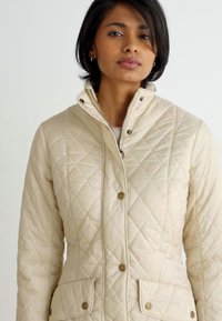 Casaco acolchoado bege claro com gola alta, botões de pressão e dois bolsos frontais com abas. Textura suave e ligeiramente brilhante.