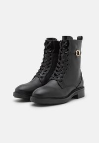 Stivaletti alla caviglia in pelle nera con chiusura frontale con lacci, punta tonda, tacco a blocco e dettagli lucidi. Presentano un logo sul lato esterno.