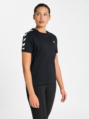 Kvinde stående let til venstre, iført sort T-shirt med hvide chevron-striber på ærmerne og sorte leggings, på ensfarvet hvid baggrund.