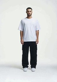 Jovem de pé, de frente, usando uma t-shirt oversized branca, calças cargo pretas e sapatilhas brancas, sobre um fundo claro e sem padrões.