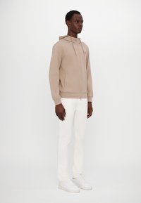 Sudadera beige con cremallera en tejido suave, presenta una capucha, dos bolsillos laterales y puños acanalados. Combinada con pantalones ajustados blancos y zapatillas blancas.