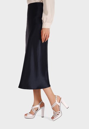 Gonna midi nera in satin abbinata a sandali bianchi con tacco alto, caratterizzati da un design a incrocio e apertura sulla punta.