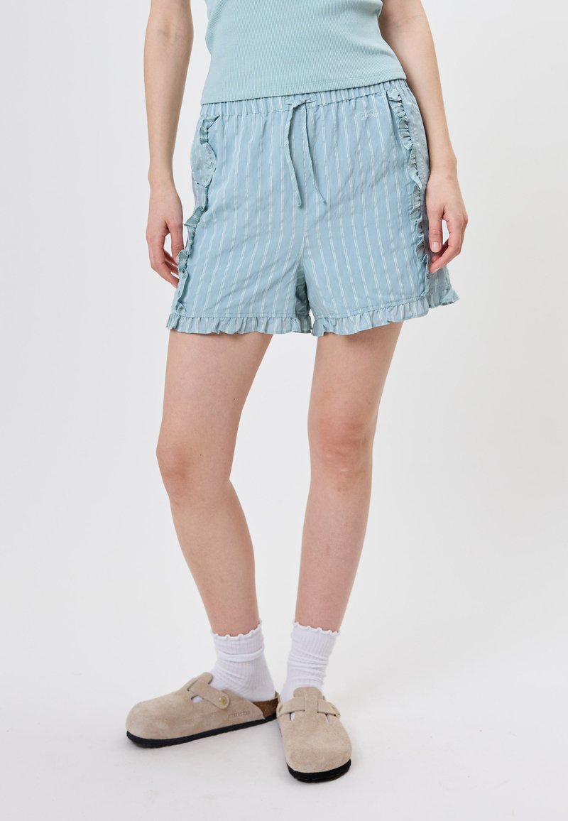 Short rayé bleu clair avec bords volantés porté par une personne en chaussettes blanches et sabots beiges à enfiler, sur un fond uni.