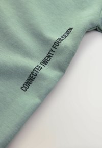 Mintunvihreä puuvillainen t-paita, jossa on lyhyet hihat ja mustalla painettu teksti "CONNECTED TWENTY FOUR SEVEN" sivusaumassa.