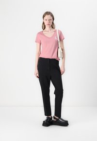 Roze V-hals T-shirt met witte logo, zwarte tailored broek en chunky zwarte instapschoenen. Model houdt een kleine beige tas vast.
