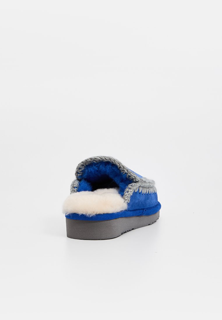 Pantofole blu in suede con bordatura intrecciata grigia, fodera in morbida pelliccia bianca e suola in gomma grigia, viste da dietro in un'ambientazione minimale.