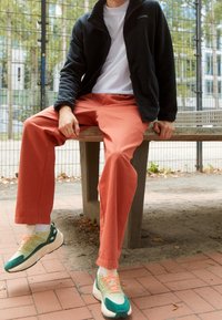 Giacca zip-up nera in pile, t-shirt bianca, chinos corallo e sneakers verdi con dettagli arancioni. Seduto su una panchina in un contesto all'aperto.