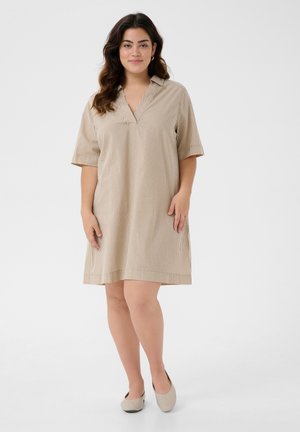 Kobieta z długimi ciemnymi włosami, ubrana w beżową, paskowaną sukienkę do kolan i beżowe baleriny, stojąca na jednolitym jasnym tle.