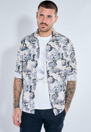 Mann mit kurzen Haaren, der ein weißes Hemd mit Blumenmuster über einem weißen T-Shirt und dunklen Hosen trägt, steht mit den Händen in den Taschen.