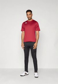 Rdeča športna majica s kratkimi rokavi in okroglim izrezom, v kombinaciji s črnimi ožjimi hlačami in belimi športnimi čevlji, z logotipom na majici.