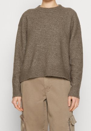 Person iført en brun uldtrøje og beige cargobukser med lynlåslommer, stående mod en ensfarvet lys baggrund.