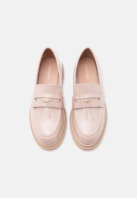 Paire de mocassins en cuir verni beige avec bouts ronds, bride décorative et emblème doré en forme de cœur, sur un fond clair.
