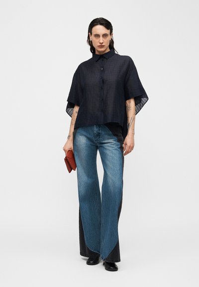 Άτομο που φοράει μια φαρδιά μαύρη πουκαμίσα με κουμπιά, μπλε τζιν παντελόνι με φαρδιά πόδια και μαύρες πλαϊνές πάνελ, μαύρα παπούτσια, κρατώντας ένα μικρό κόκκινο φάκελο.