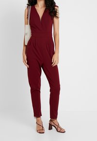 Burgundowy kombinezon z głębokim dekoltem w kształcie litery V, bez rękawów, dopasowaną talią i zwężanymi nogawkami, zestawiony z fakturowaną, przezroczystą torebką.