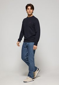 Marineblauwe sweatshirt met geribbelde manchetten en halslijn, met een klein rood logo. Gepaard met lichtblauwe straight-leg jeans en beige sneakers.