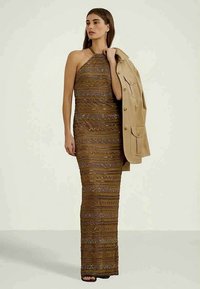 Γυναίκα φορώντας ένα μακρύ, μοτίβο, αμάνικο καφέ φόρεμα, κρατώντας ένα μπεζ μπουφάν στον έναν ώμο, καθώς στέκεται μπροστά από ένα απλό φωτεινό φόντο.