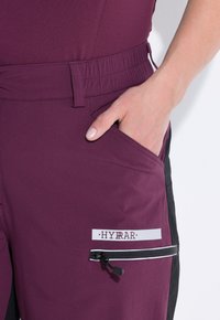 Burgundské sportovní kalhoty s kontrastním černým bočním panelem, elastickým pasem, boční kapsou a kapsou na zip s bílým akcentem.