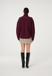 Burgunderroter oversized gestrickter Pullover mit hohem Kragen, kombiniert mit einem beige texturierten Minirock und schwarzen kniehohen Stiefeln mit Absatz.