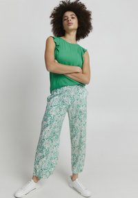 Blusa verde senza maniche con dettagli arricciati sulle spalle, abbinata a pantaloni bianchi larghi con un motivo floreale verde, indossati con sneakers bianche.