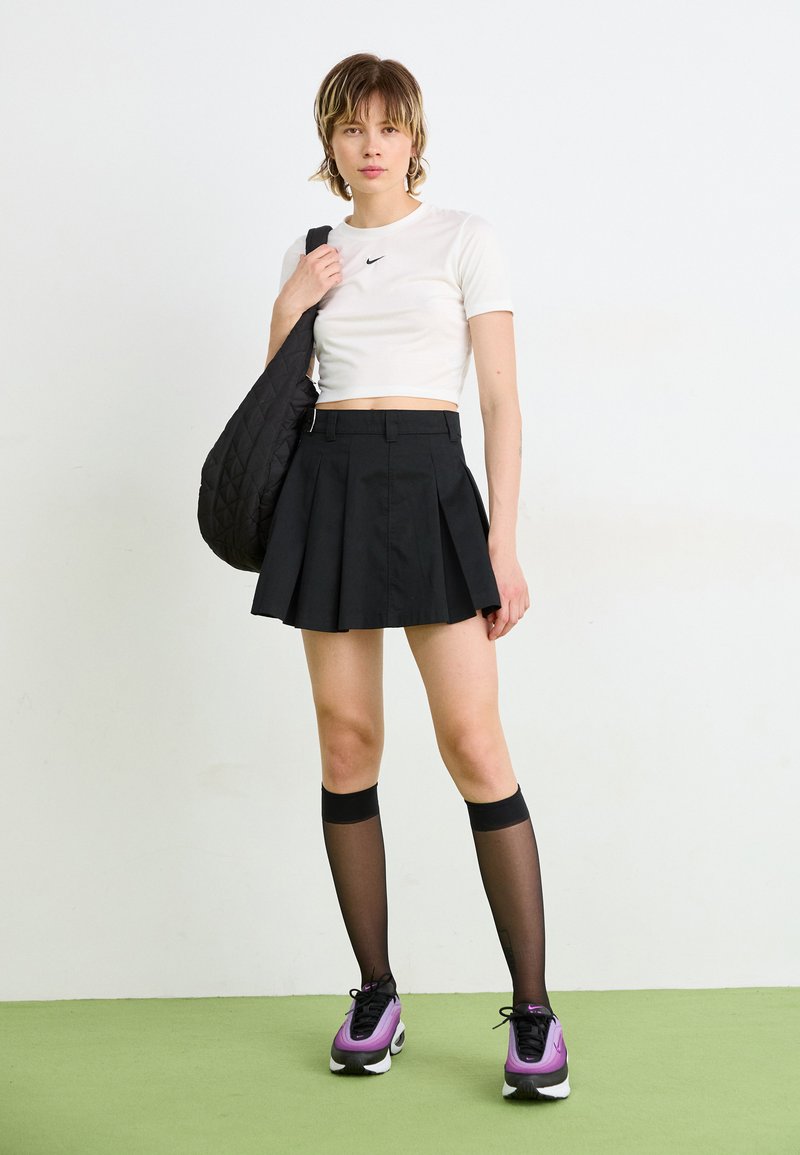 T-shirt bianco cropped con logo Nike nero, gonna nera plissettata, calze nere al ginocchio e sneakers viola/nere, mentre porta una borsa nera trapuntata.