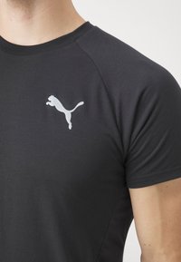Melnā T-kreklā ar īsām piedurknēm, ar atstarojošu baltu Puma logotipu kreisajā krūtīs. Izgatavots no gludas, elastīgas auduma.
