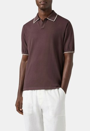 Uomo che indossa una polo marrone scuro a maniche corte con bordino bianco su colletto e polsini, abbinata a pantaloni bianchi con tasche frontali.