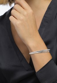 Silbermetallarmband mit einer gedrehten Gestaltung, das eine unregelmäßige Textur aufweist. Wird an einer Hand mit neutralem Hautton getragen, was einen Kontrast zu einem dunklen Oberteil bildet.