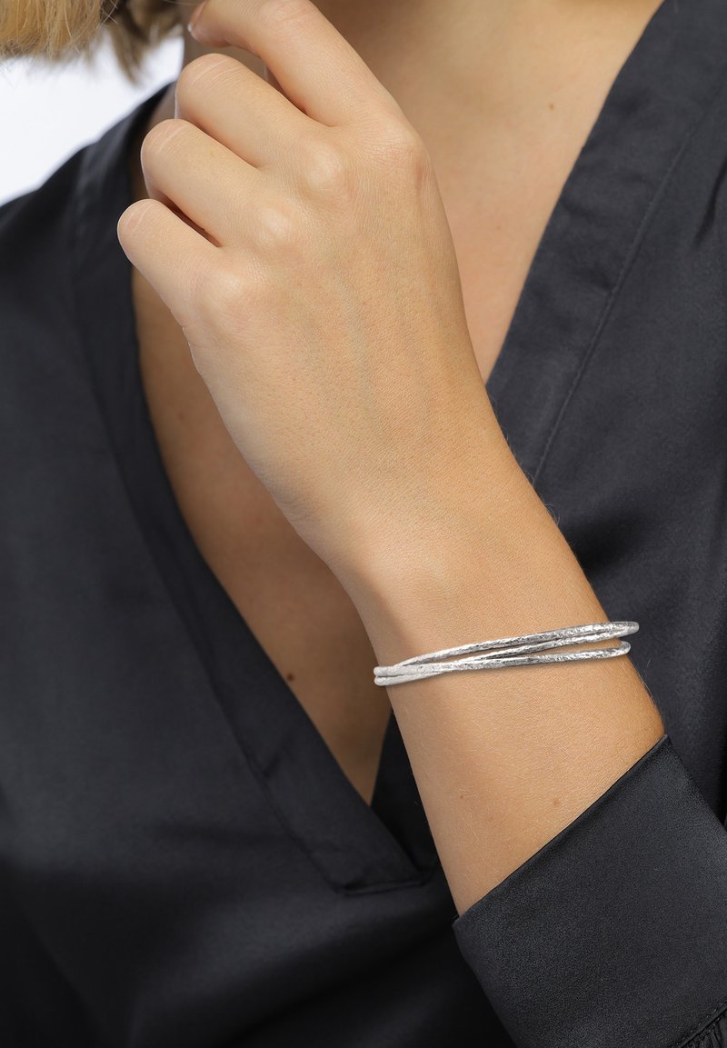 Silbermetallarmband mit einer gedrehten Gestaltung, das eine unregelmäßige Textur aufweist. Wird an einer Hand mit neutralem Hautton getragen, was einen Kontrast zu einem dunklen Oberteil bildet.