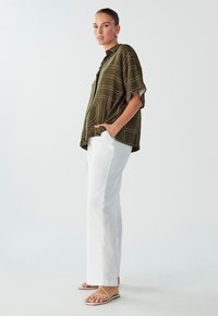 Camicia verde oliva con stampa geometrica bianca, maniche corte e vestibilità rilassata, abbinata a pantaloni bianchi a gamba dritta e sandali nudi.