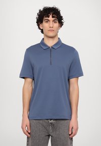 Korte ermer, blå polo skjorte med glidelås i kragen. Laget av glatt stoff. Kombinert med lysegrå, falmede jeans.