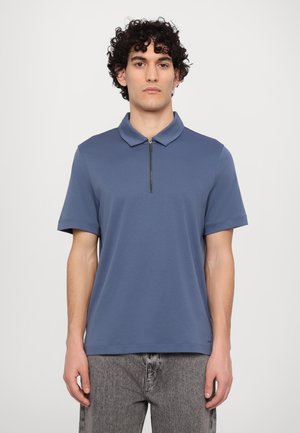 Polo shirt bleu à manches courtes avec un col zippé. Fabriqué en tissu lisse. Associé à un jean délavé gris clair.
