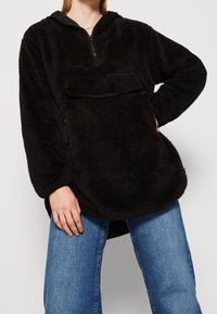 Donna che indossa un pullover in fleece nero con tasca frontale e mezzi zip, abbinato a jeans di denim blu, in posa con una mano sul fianco.