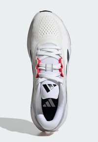 Chaussure de sport blanche avec une tige tricotée respirante, des accents rouges, un logo noir et un amorti visible dans la semelle. Présente un système de fermeture à lacets.