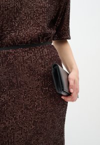 Melna ādas somiņa, ko tur rokā, saskaņota ar mirdzošu brūnu kleitu, kas izceļas ar teksturētu sekvīnu rakstu, maigu audumu un šauru jostu.
