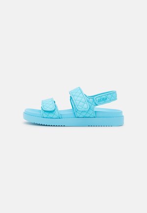 Lyseblå quiltet sandal med to justerbare stropper over foden og en rundt om anklen, flad sål med rillede bund.