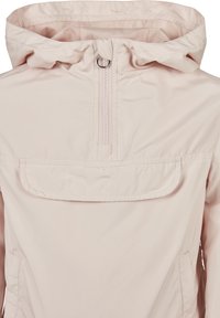Veste à capuche rose clair avec un demi-zip à l'avant, des poignets élastiques et une grande poche frontale avec rabat. Tissu lisse et résistant à l'eau.