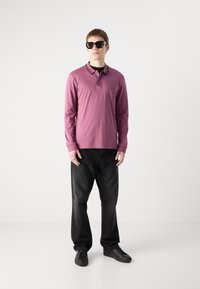 Camisa polo de manga longa em roxo suave com colarinho e detalhes em preto, combinada com calças de ganga largas pretas e ténis pretos.