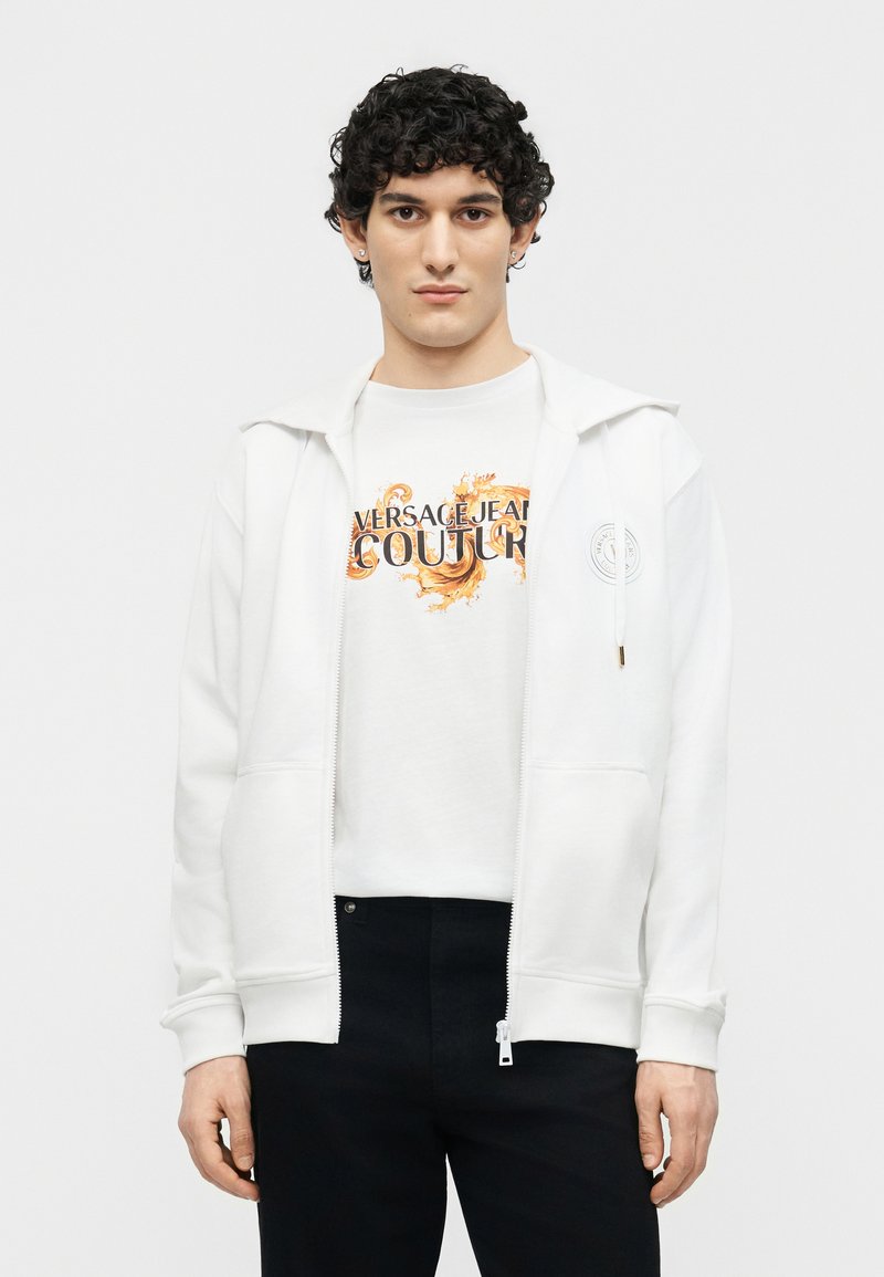 Νέος άνδρας με σγουρά μαλλιά που φοράει λευκή ζακέτα με φερμουάρ πάνω από φανέλα Versace Jeans Couture και μαύρο παντελόνι, κοιτάζοντας μπροστά.