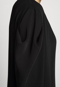 Svart långärmad plagg med en strukturerad silhuett, som har en rund halsringning och mjukt puffade ärmar. Tyget ser slätt ut och faller elegant.