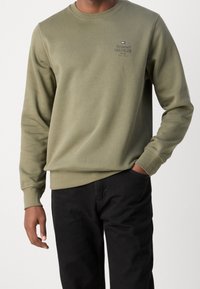 Felpa verde oliva con scollo a girocollo, polsini e orlo a coste. Presenta una piccola stampa del logo sul petto. Abbinata a pantaloni neri.