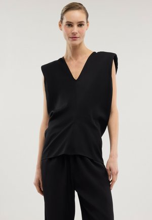 Frau mit hellem Teint und hochgestecktem Haar, trägt ein ärmelloses schwarzes V-Ausschnitt-Oberteil mit gepolsterten Schultern und schwarze Hosen vor einem schlichten, hellen Hintergrund.