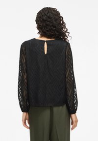 Black blouse with sheer, patterned fabric featuring wave-like textures. Keyhole detail at the back; long, slightly puffed sleeves.
