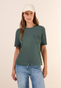 T-shirt vert olive à manches courtes, avec une poche texturée et un ourlet à cordon. Associé à un jean bleu clair.