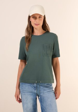 Camiseta verde oliva de manga corta, con un bolsillo texturizado y un dobladillo con cordón. Combinada con unos vaqueros azul claro.